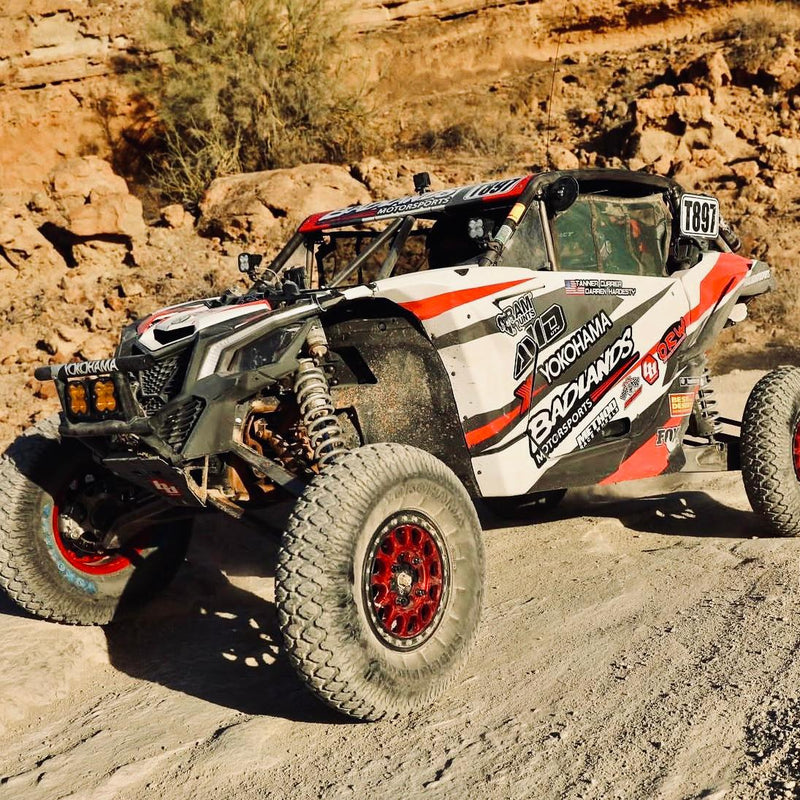 Red, white, and black Maverick X3 race vehicle on rocky terrain with front suspension visible